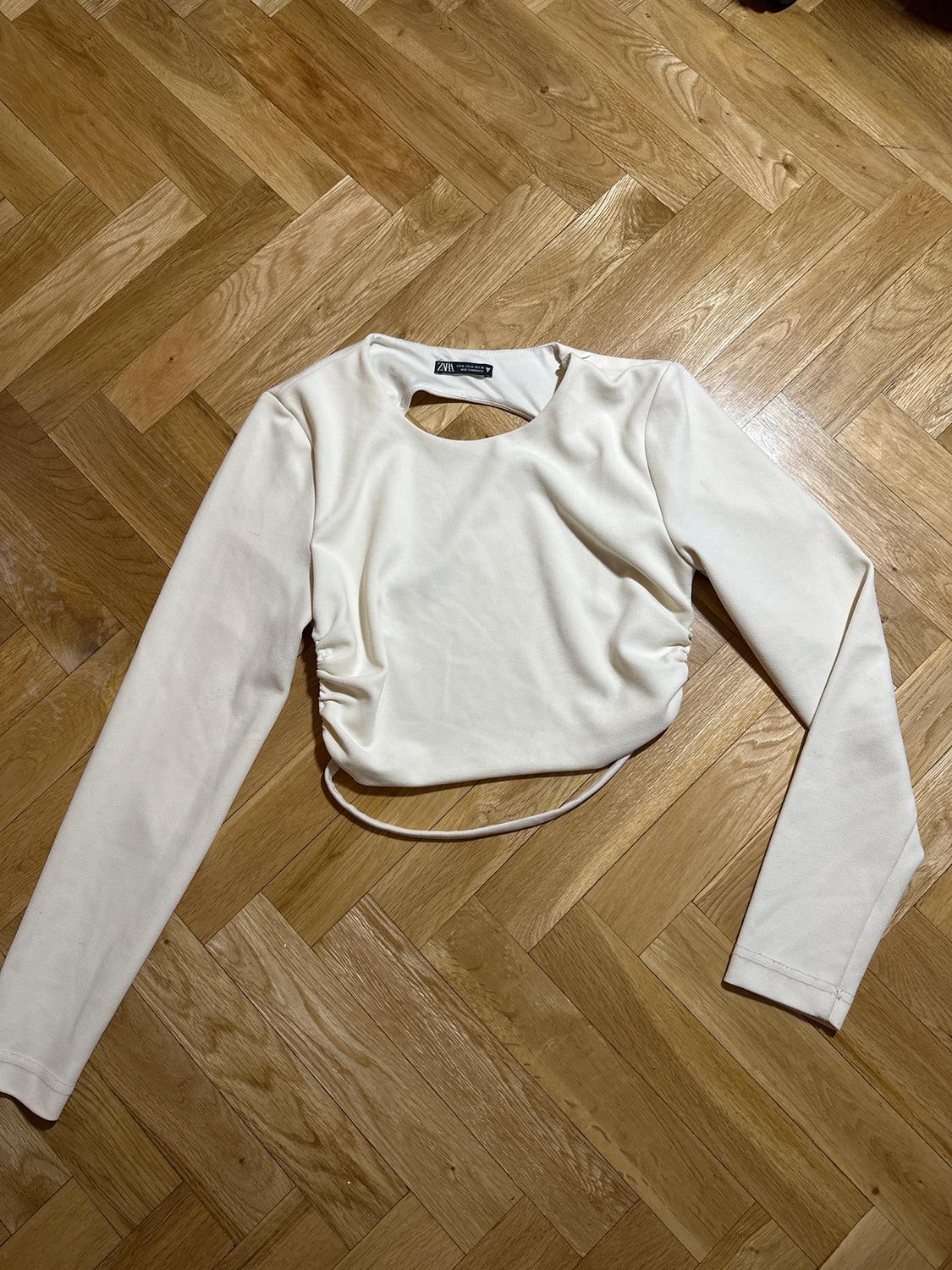 White long-sleeved T-shirt with a chest opening, on a light wooden floor. Visible label: "Zara".