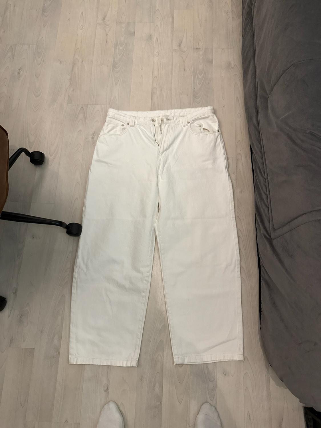 Wide white jeans on a light wooden floor, between a chair and a grey sofa. At the bottom, feet wearing socks are visible.