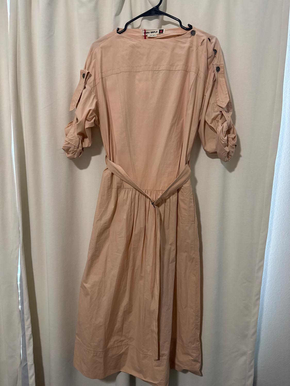 Beige short-sleeved dress with buttons on the shoulders, hanging on a black hanger in front of a white curtain.