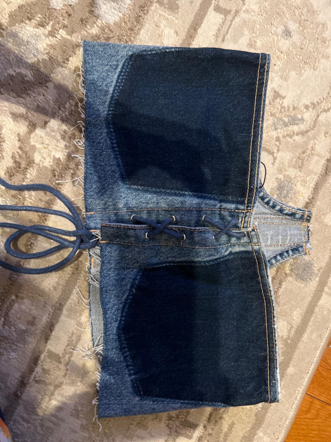 Blue denim corset with dark pockets, adjustable laces, and frayed edges, placed on a beige rug with a geometric design.