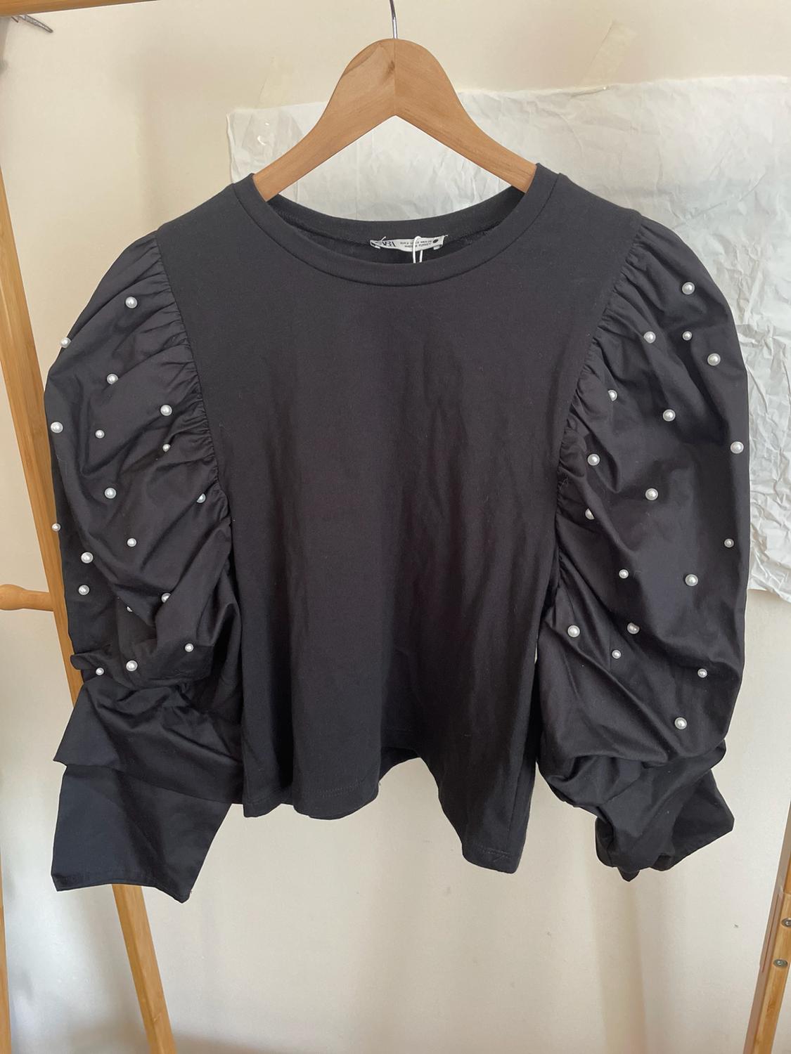 Black long-sleeved top with puffed sleeves and white pearl detailing, hanging on a wooden hanger against a light background.