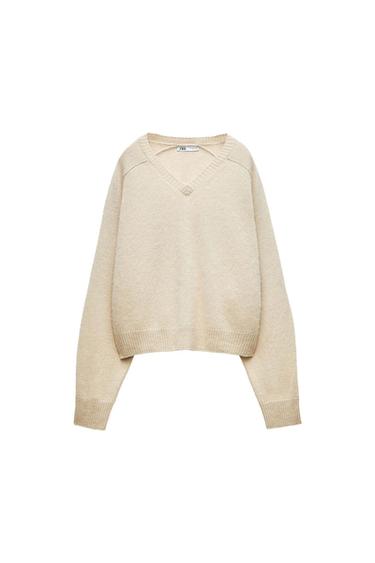 Beige knitted V-neck jumper with long sleeves, hanging against a white background. Label visible on the collar.