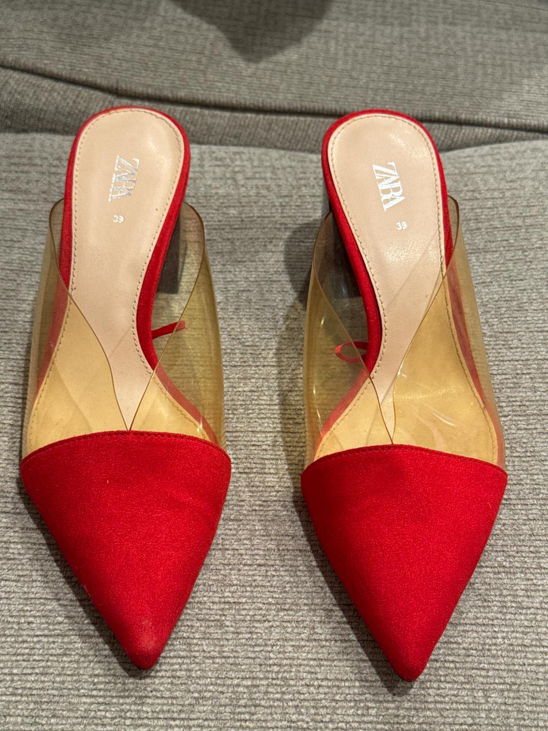 Red-toed high-heeled shoes with transparent details on a grey surface. Visible text: "ZARA 39".