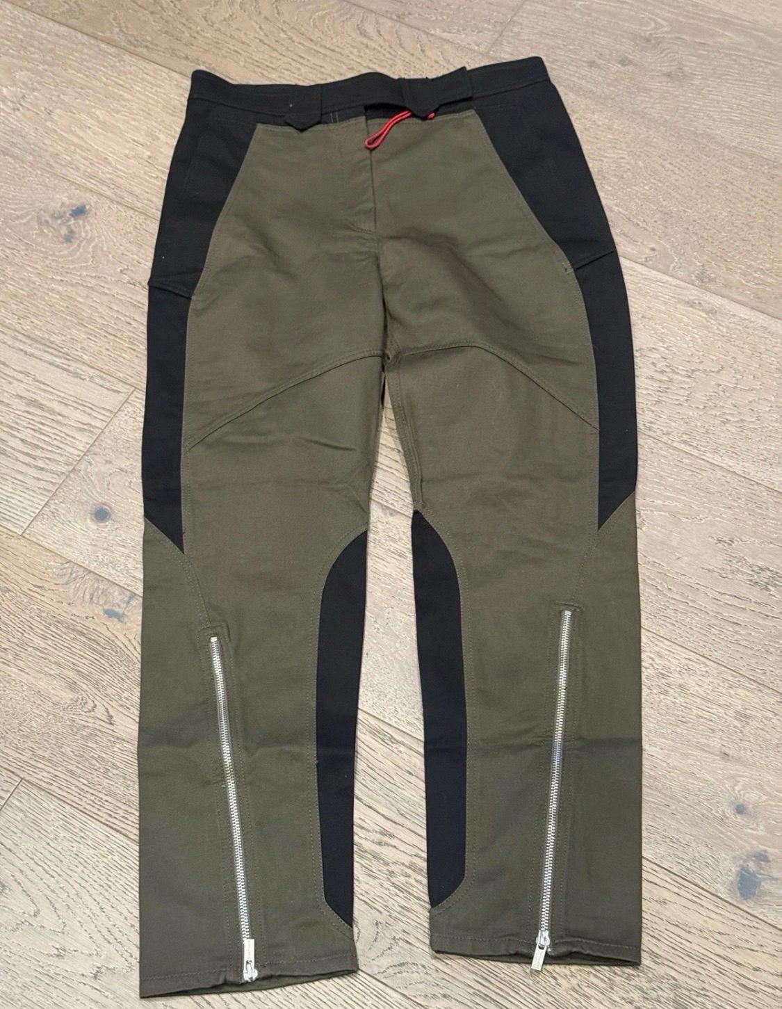 Olive green technical-style pants with black panels and zippers at the hems, on a light wood floor.