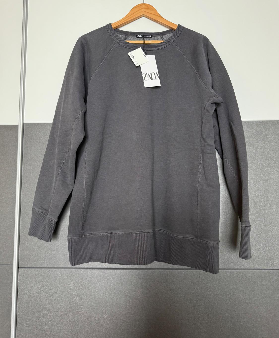 Grey long-sleeved sweatshirt hanging on a wooden hanger. Visible label with the text "ZARA". Grey and white background.