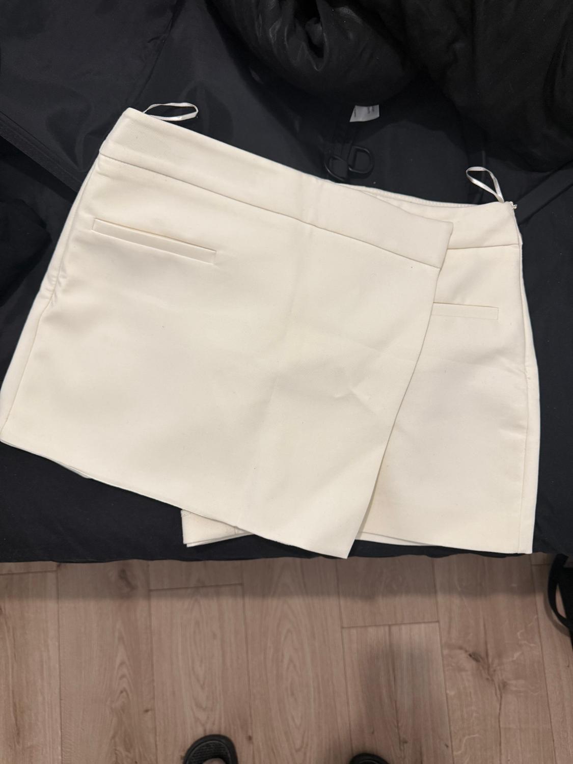White mini skirt on a black background, with front pockets and visible stitching details. Light wooden floor in the setting.