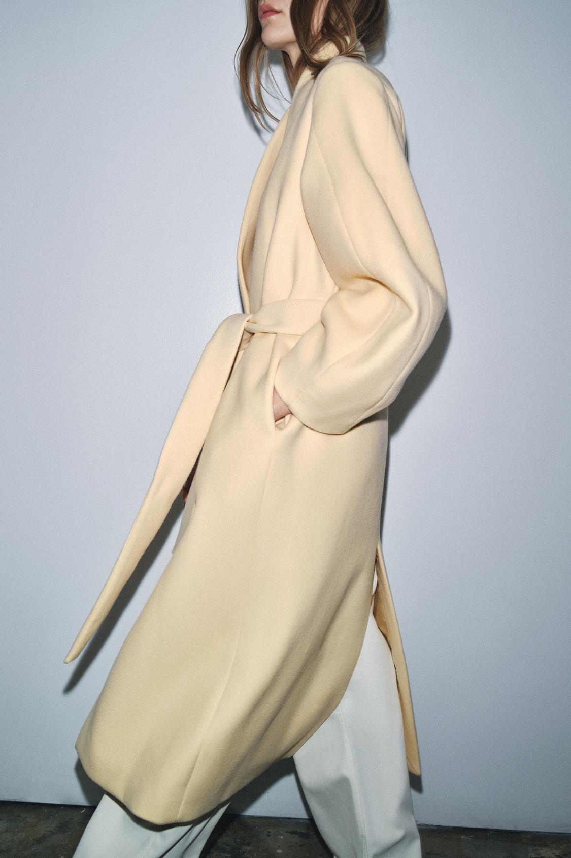 Oversize vanilijev komplet s plaščem z okroglim ovratnikom, dolgimi rokavi in nastavljivim pasom. Skriti žepi v šivu.