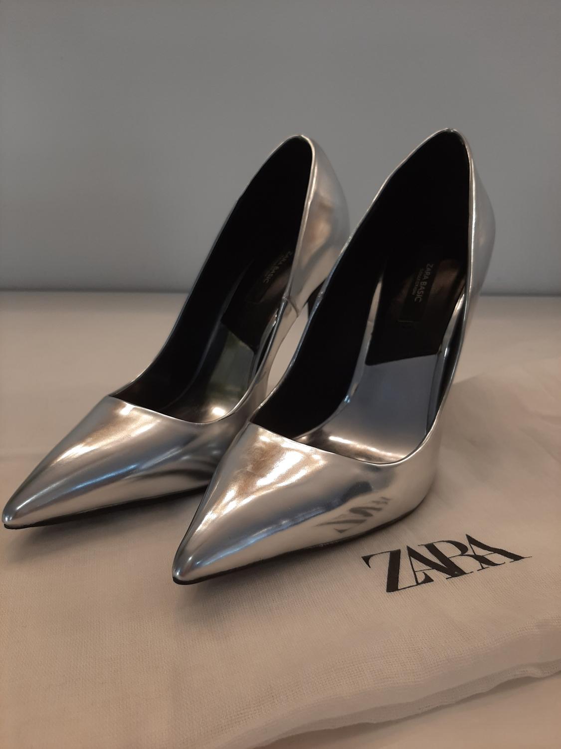 Detail of silver high-heeled shoes with a pointed toe on a bag with the text 'ZARA'.