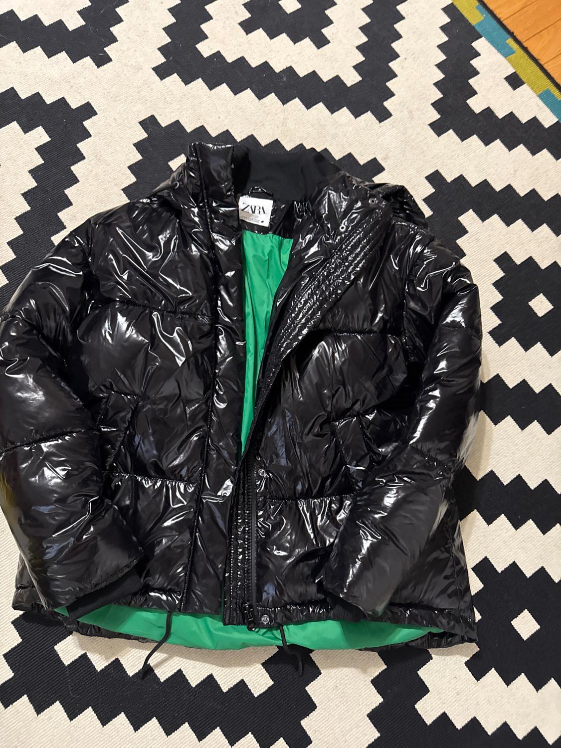 Front view of a shiny black padded jacket with a hood and zip, green interior.