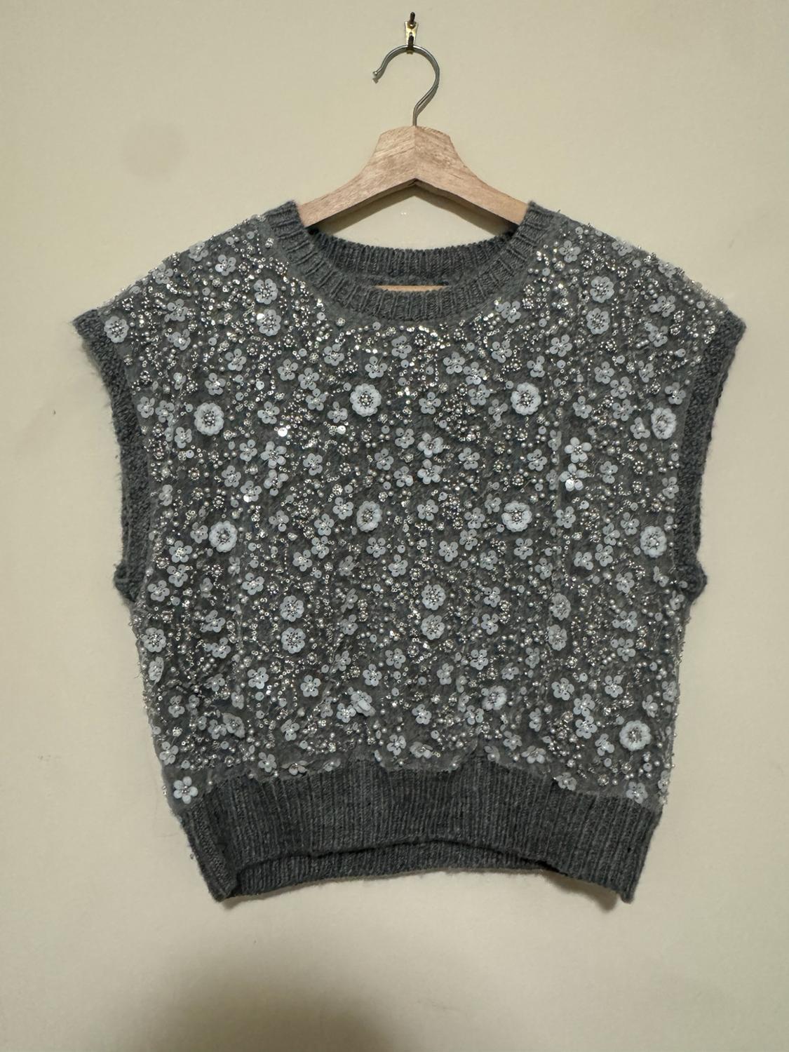 Grey sleeveless knit top hanging on a wooden hanger against a light background, adorned with sequins, beads, and white flowers.