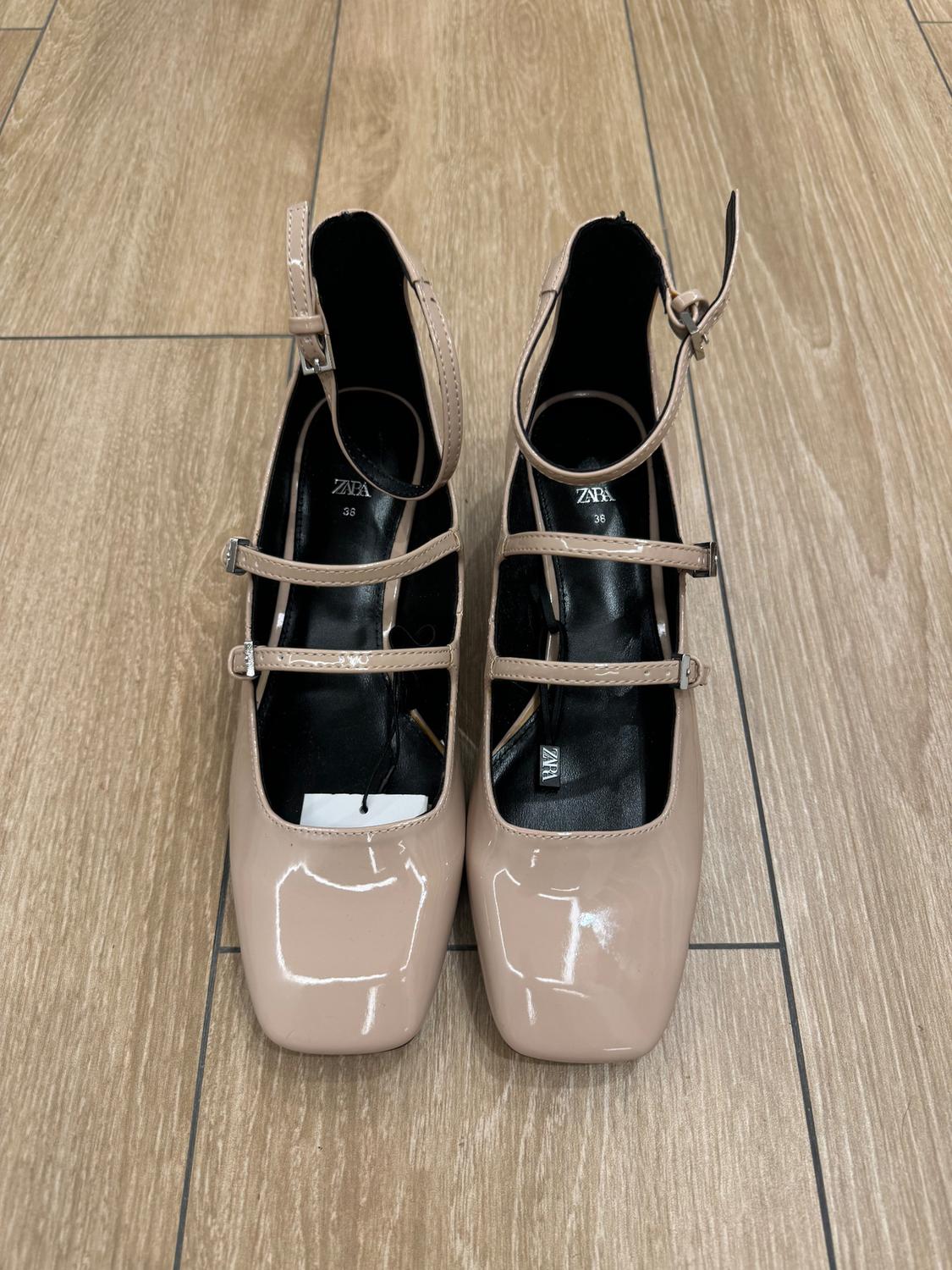 Beige patent leather shoes with straps and buckles, viewed from above on a wooden floor. Visible text: "ZARA 36".
