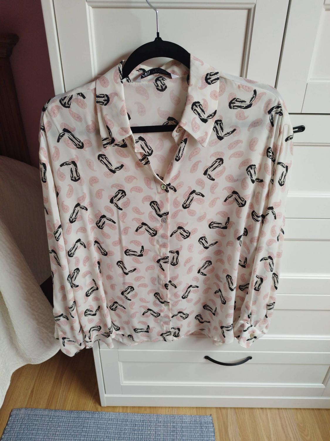 White shirt with a black boot and pink motif print, hanging on a black hanger in front of a white wardrobe in a bedroom.