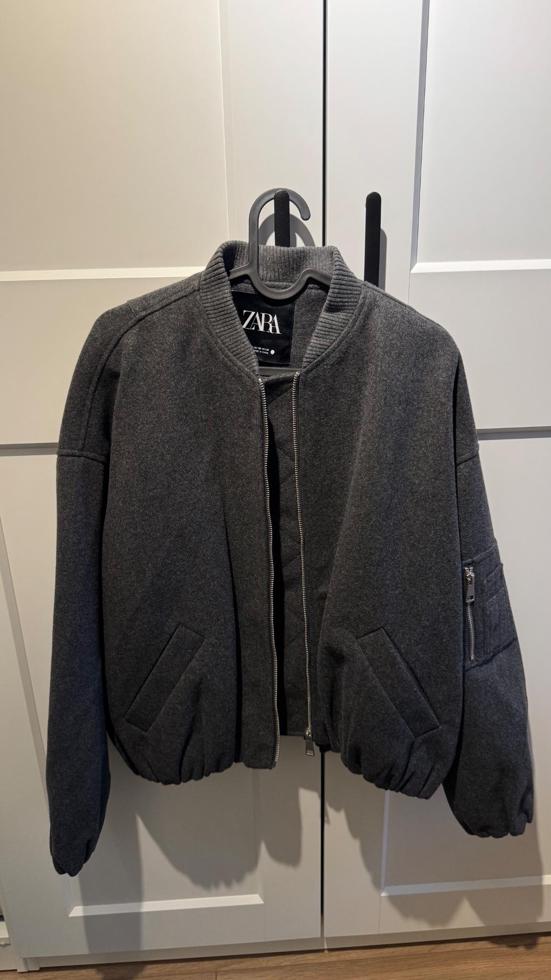 Grey Zara jacket hanging on a black hanger in front of a white wardrobe. Zip-up design with side pockets.