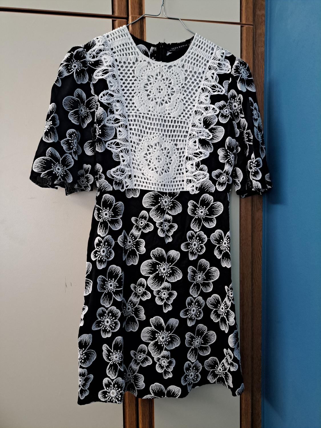 Black dress with a white floral print and lace detail on the chest, hanging on a hanger against a blue and beige background.