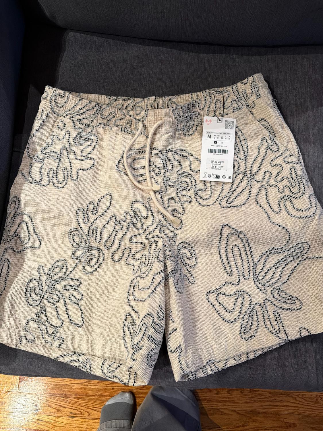 Beige shorts with a dark line print, an elastic waistband with a drawstring, and a hanging price tag, on a gray background.