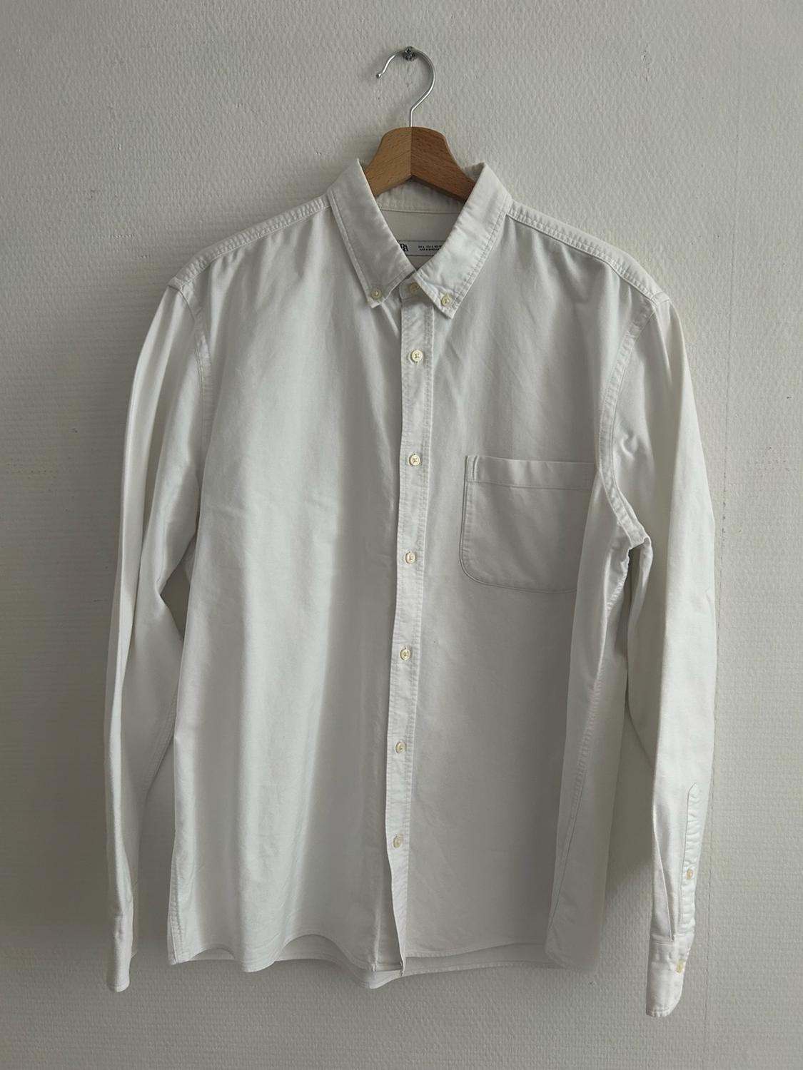 Long-sleeved white button-up shirt with a pocket, hanging on a wooden hanger against a white wall. Minimalist style.