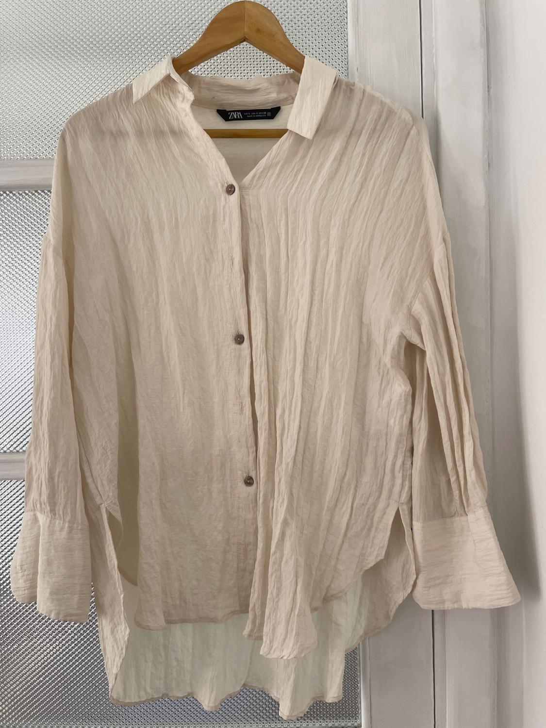Beige shirt made of crinkled fabric hanging on a wooden hanger. Door background with textured glass. Visible label: "Zara".