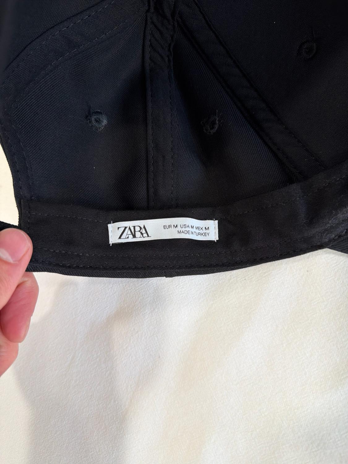 Hand holding the inside of a black cap. Label with text: ZARA EUR M USA M MEX M MADE IN TURKEY.
