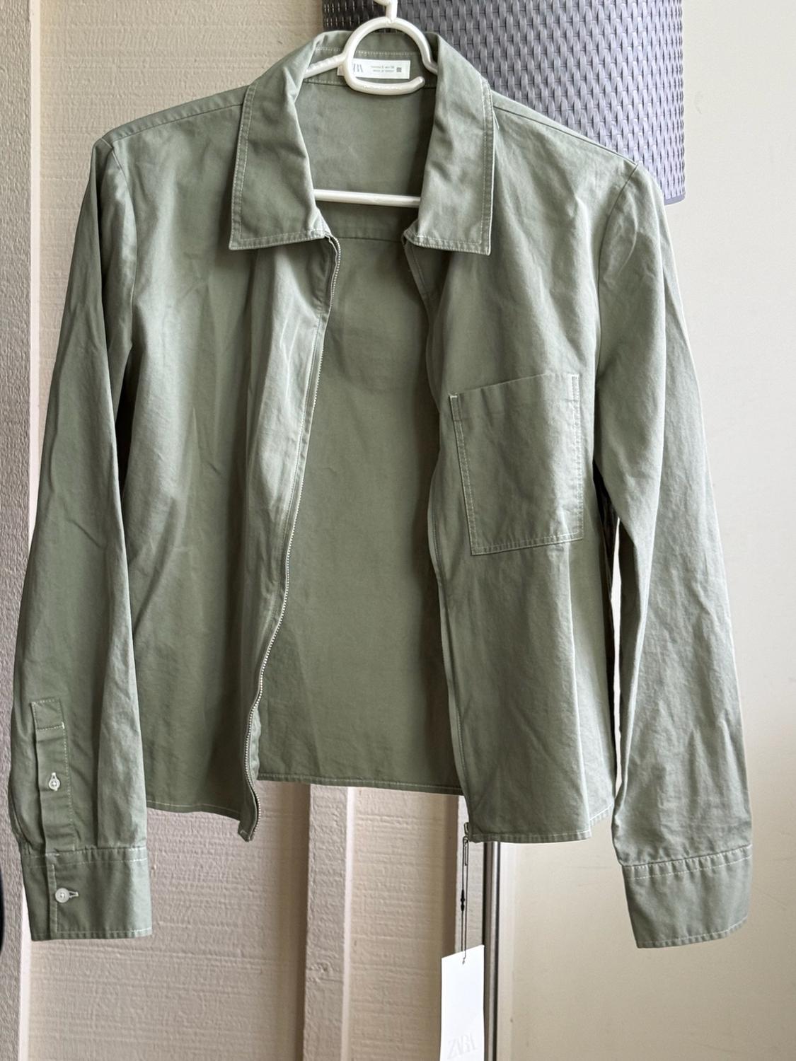 Olive-green jacket with a zip and pocket, hanging on a white hanger against a neutral background. Label with the text ZARA visible.
