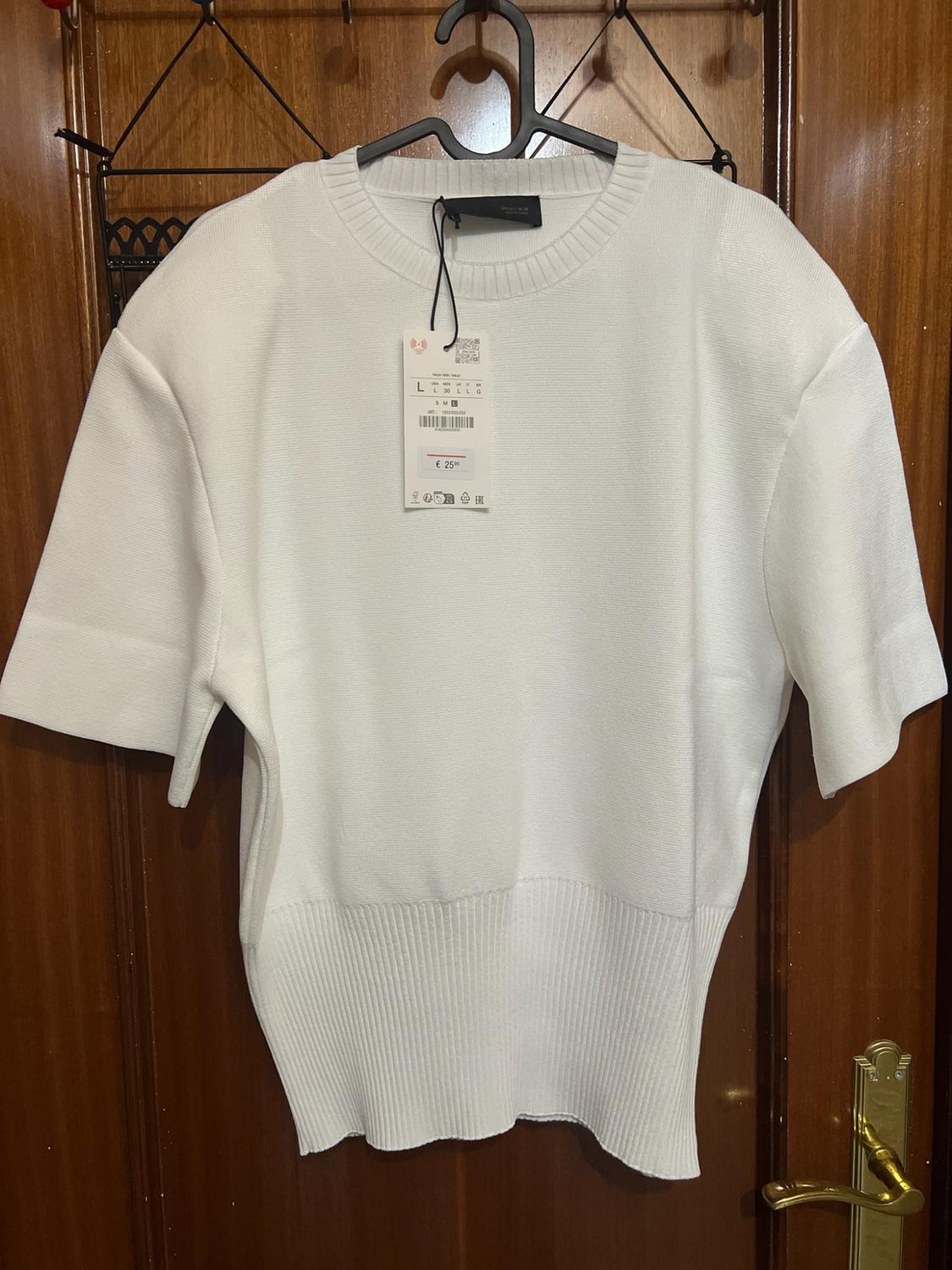 White polka-dot top with short sleeves and a ribbed hem, hanging on a wooden door. Includes a label with the text “L” and “€ 25”.
