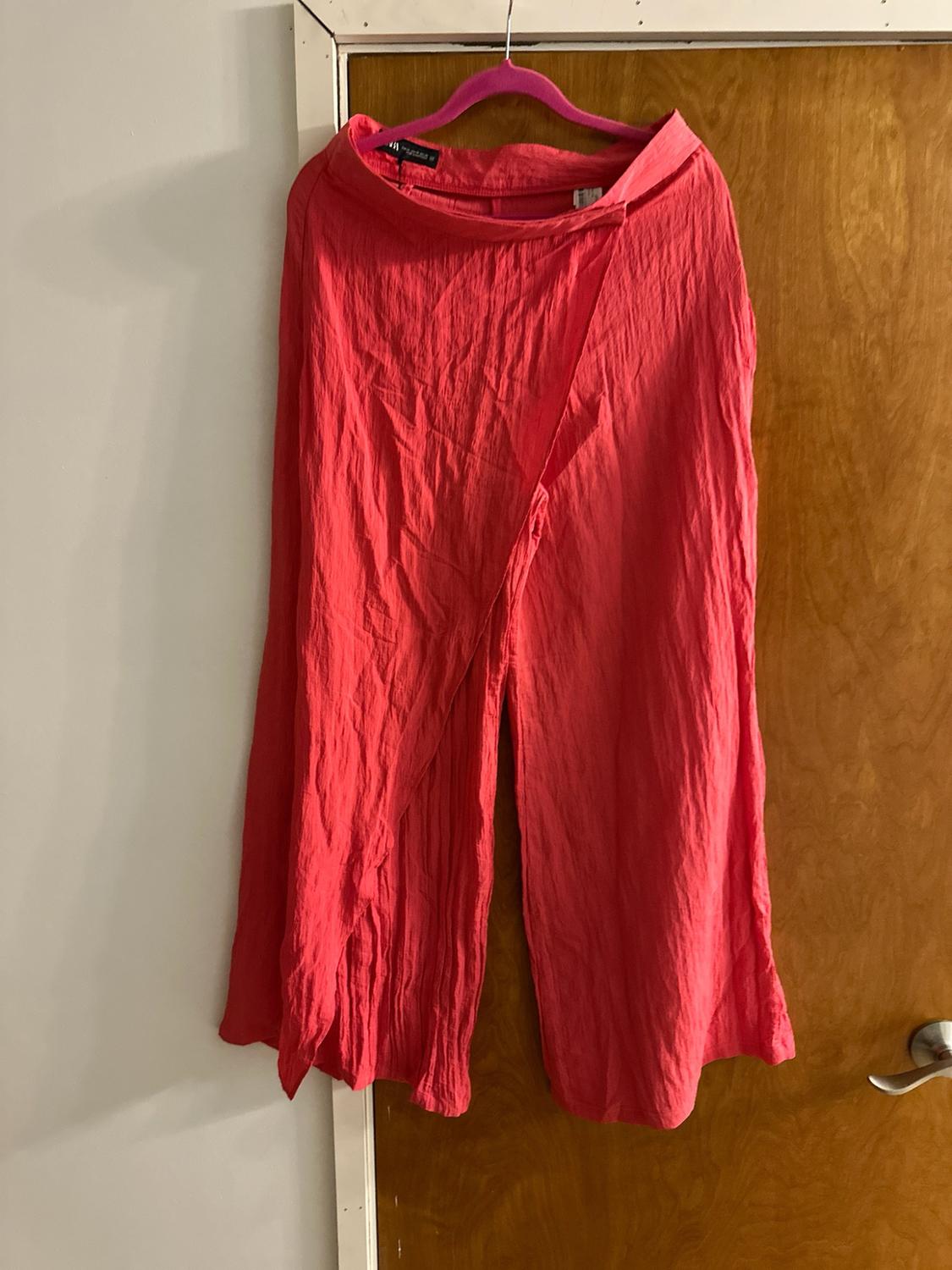 Coral pink wide-leg trousers with a wrap-over design and textured fabric, hanging on a hanger over a wooden door.