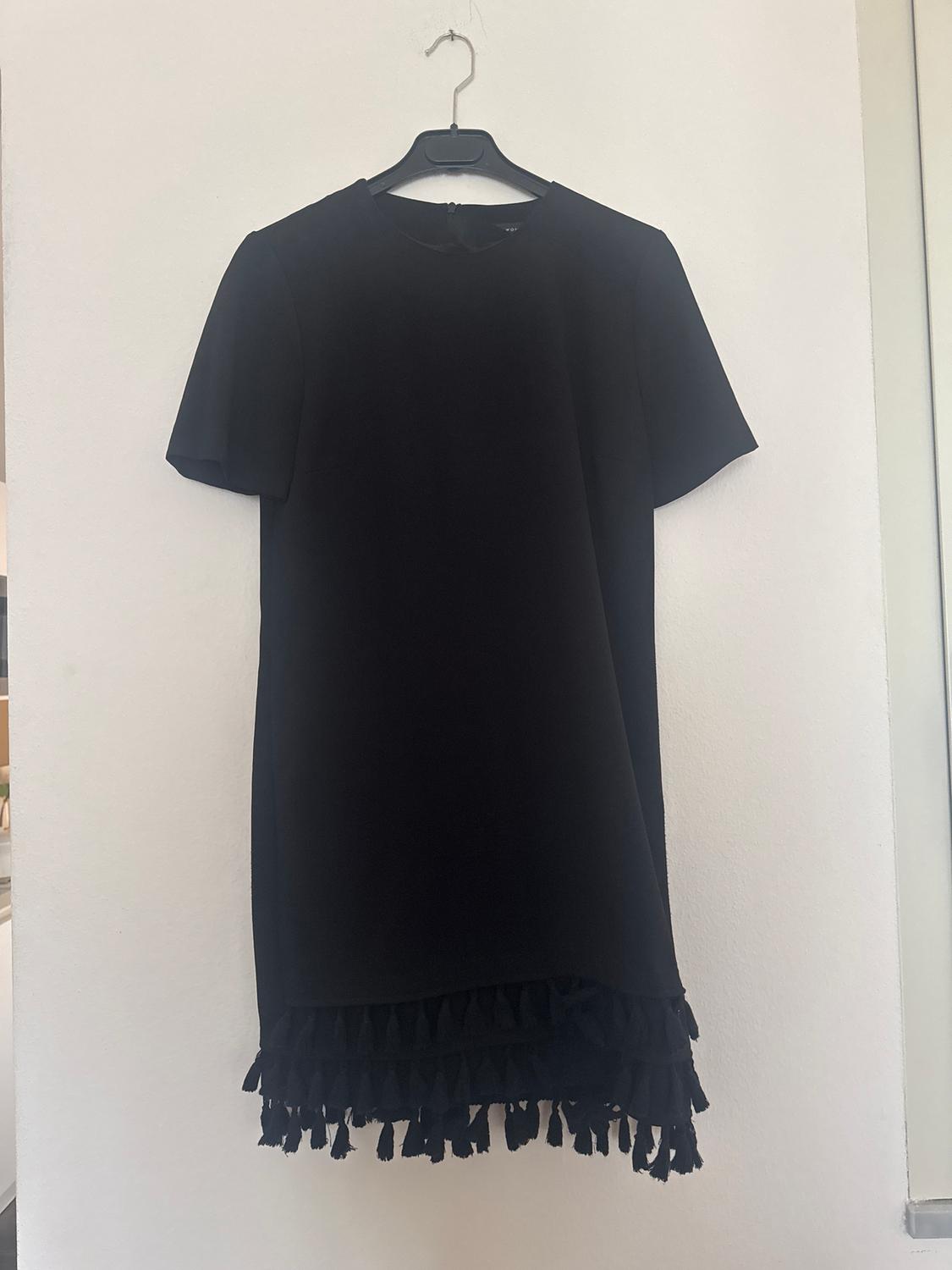 Black short-sleeved dress hanging on a hanger, with fringed detailing on the hem, on a white background.