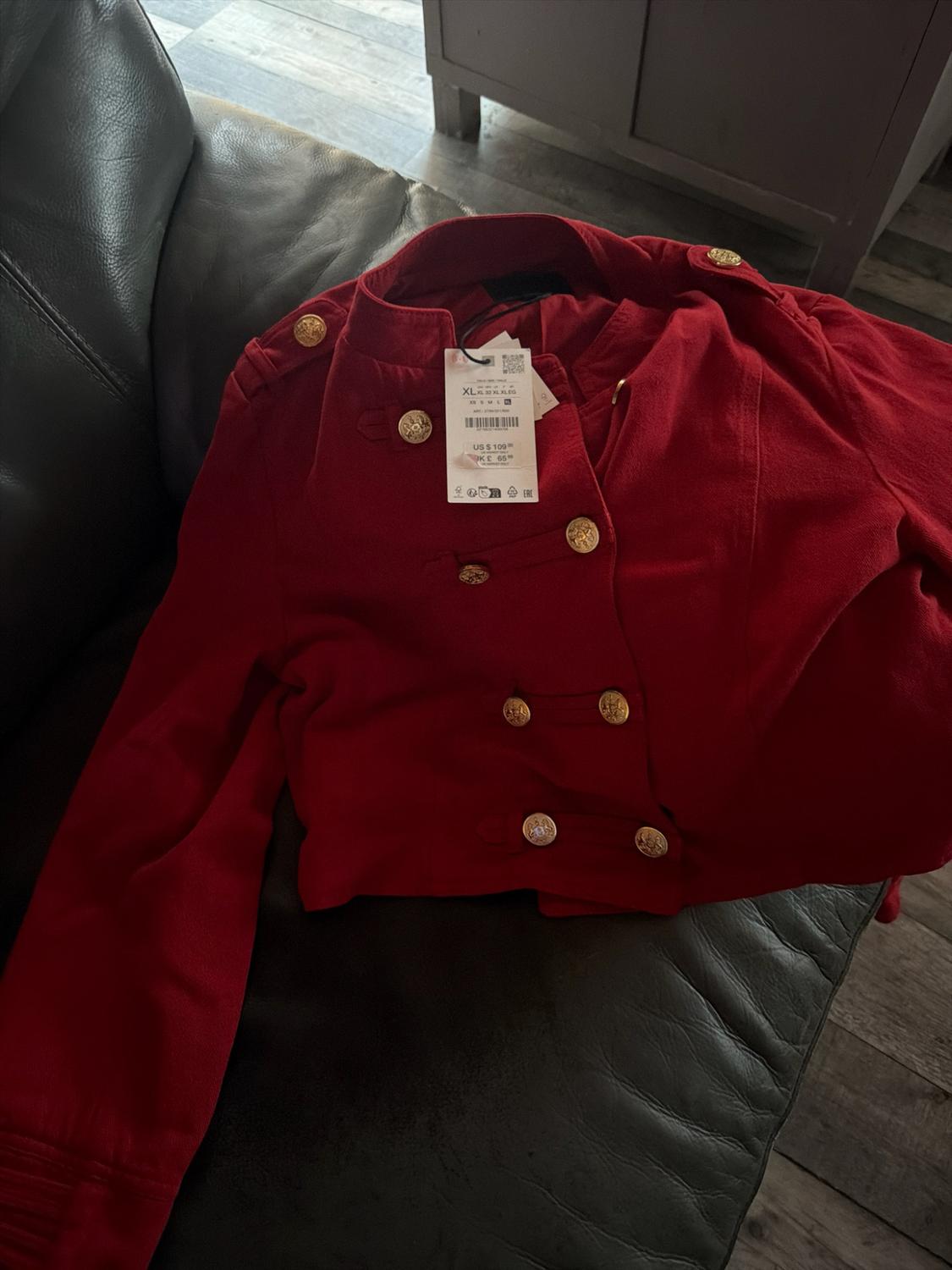 Red jacket with gold buttons on a black leather sofa. Label with text: XL US $109.00.
