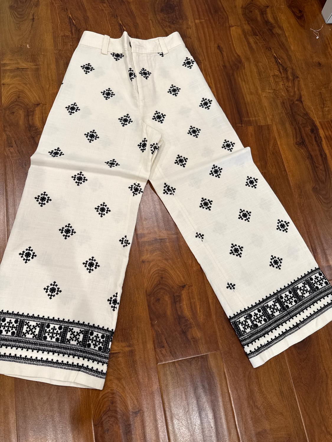 White trousers with a black geometric pattern print and embroidery on the hem, on a brown wooden floor.