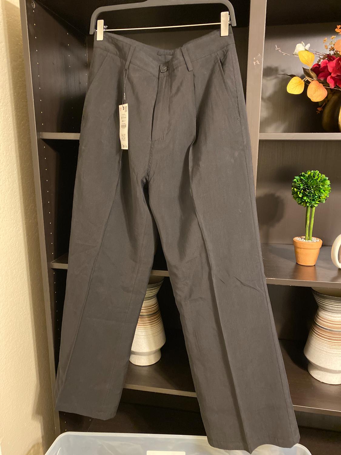 Black trousers hanging on a hanger in front of a dark shelf with plant and vase decorations. Label visible on the left side.