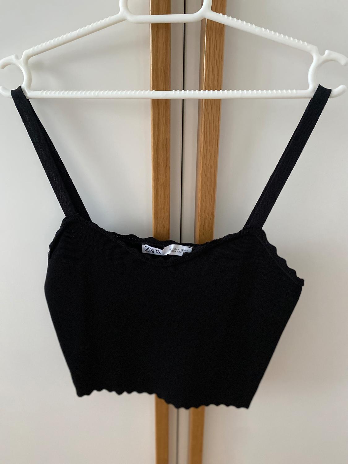 Black vest top hanging on a white hanger against a light wood wardrobe background. Label visible on the inside.