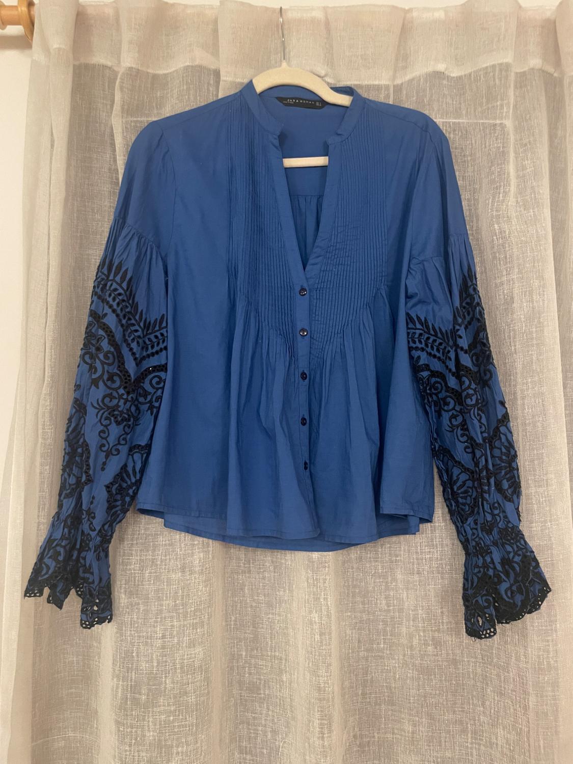 Blue blouse with long sleeves and black embroidery hanging on a hanger in front of a beige curtain.