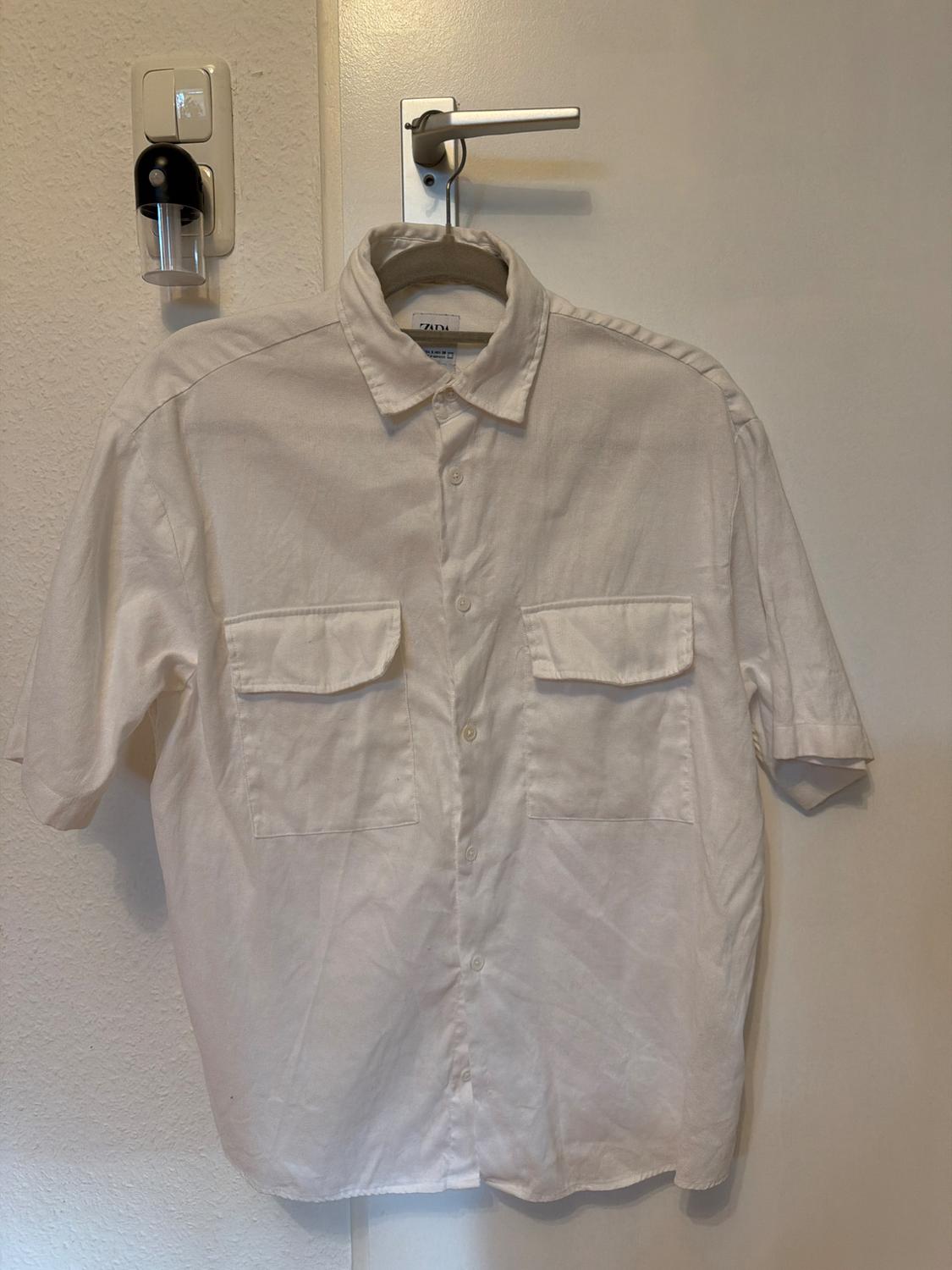 White short-sleeved shirt hanging on a white door. It has two front flap pockets and buttons. Visible label: ZARA.
