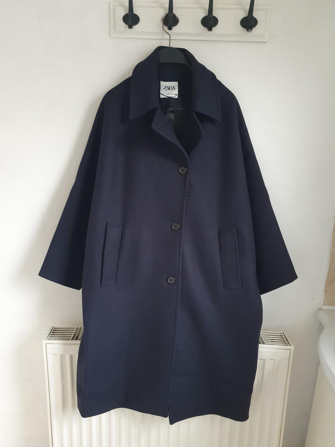 Dark blue coat hanging on a hanger with a visible Zara label, against a white background with black hooks on the wall.