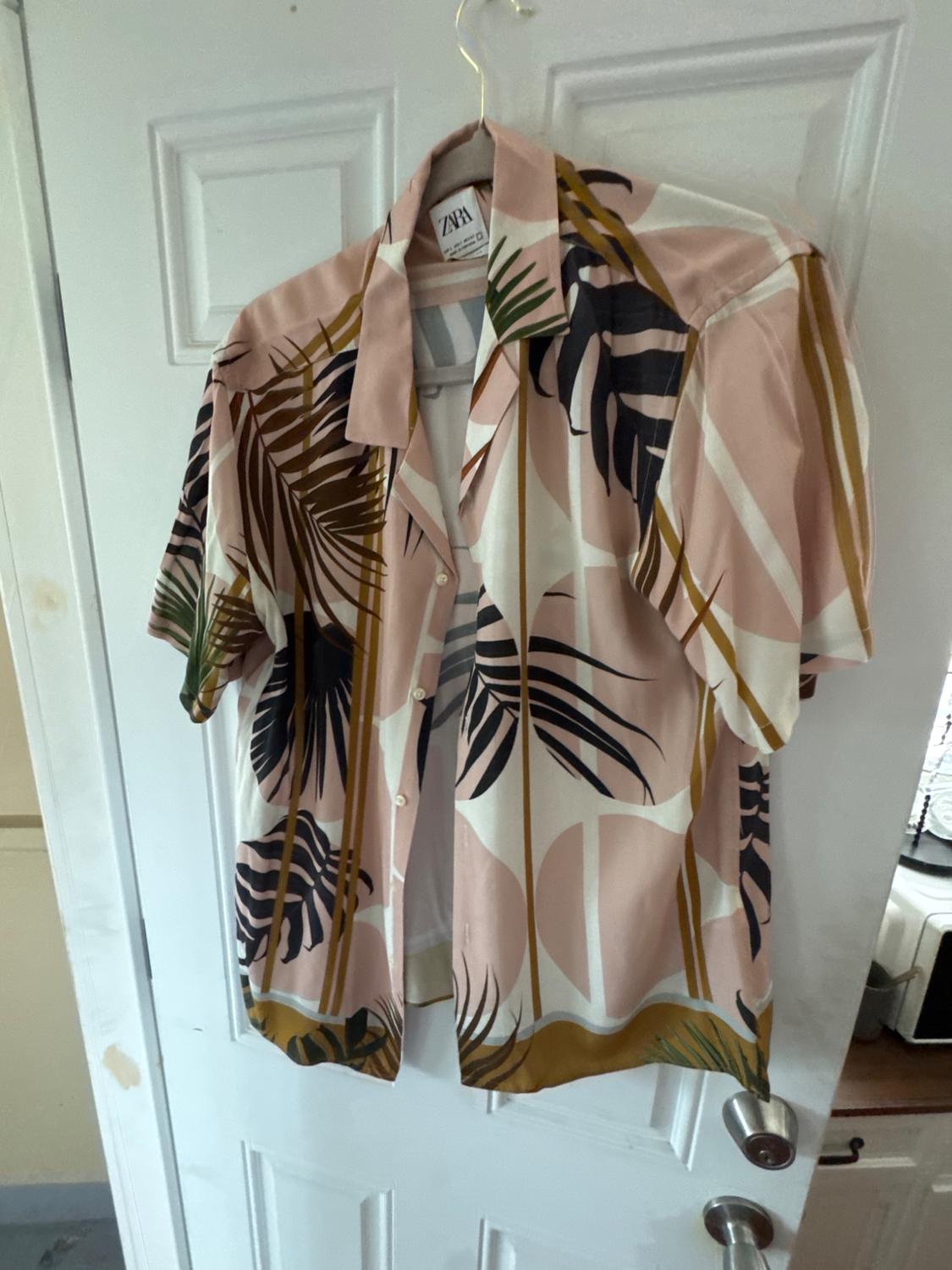 Short-sleeved shirt with tropical leaf print, hanging on a hanger over a white door. Label with the text ZARA.