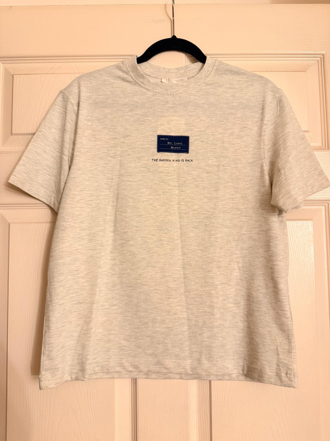 Heather gray t-shirt on a hanger over a door. Text: "SEND TO: Mr. Lawn Mower THE GARDEN KING IS BACK".