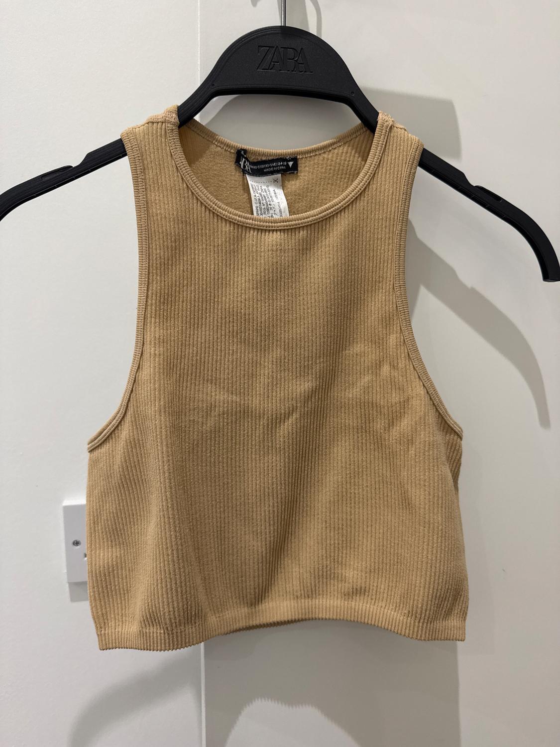Beige ribbed crop top hanging on a black hanger with visible branding. Plain white background.