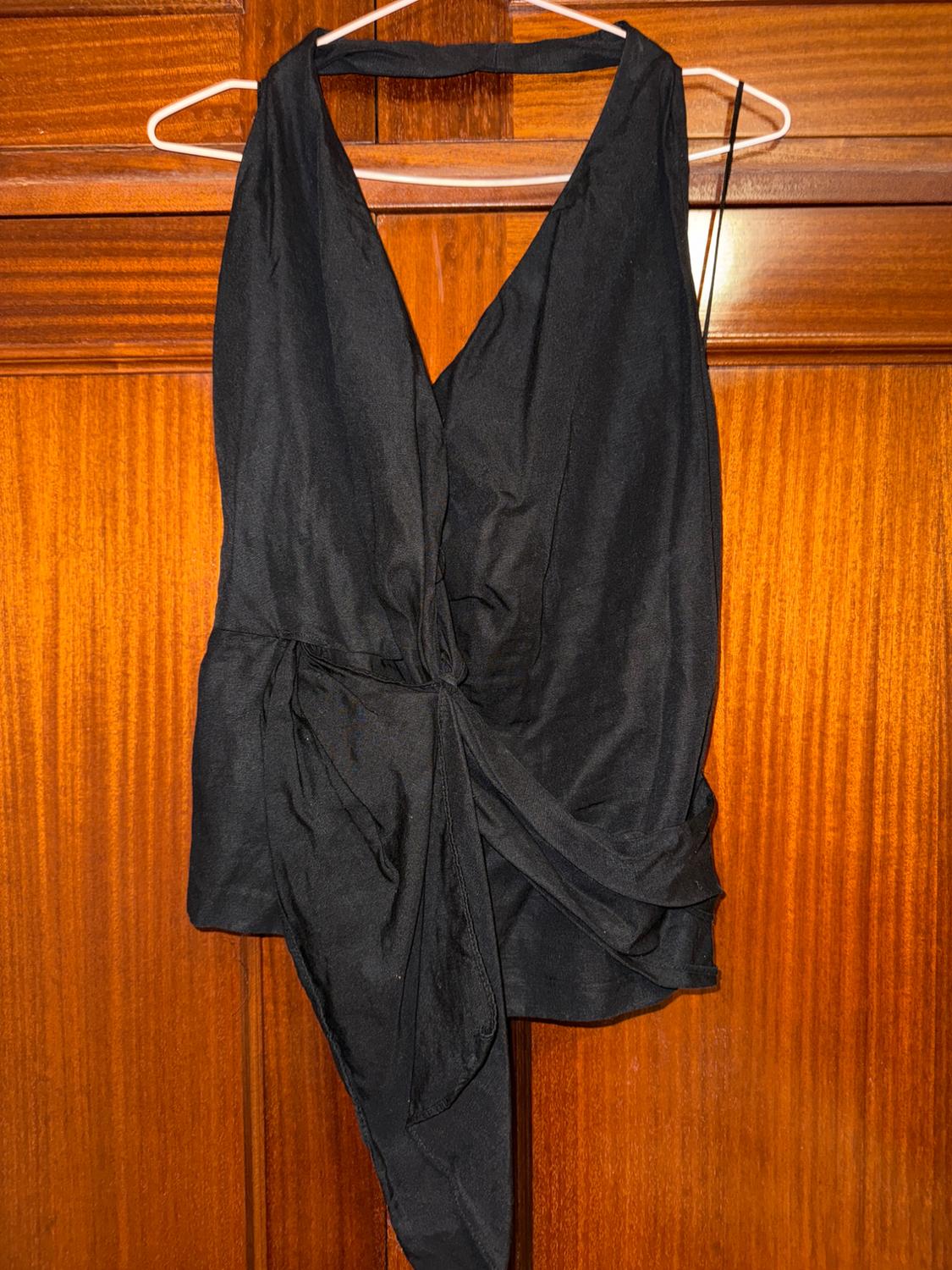 Black sleeveless blouse with a V-neck and waist tie, hanging on a white hanger over a wooden door.
