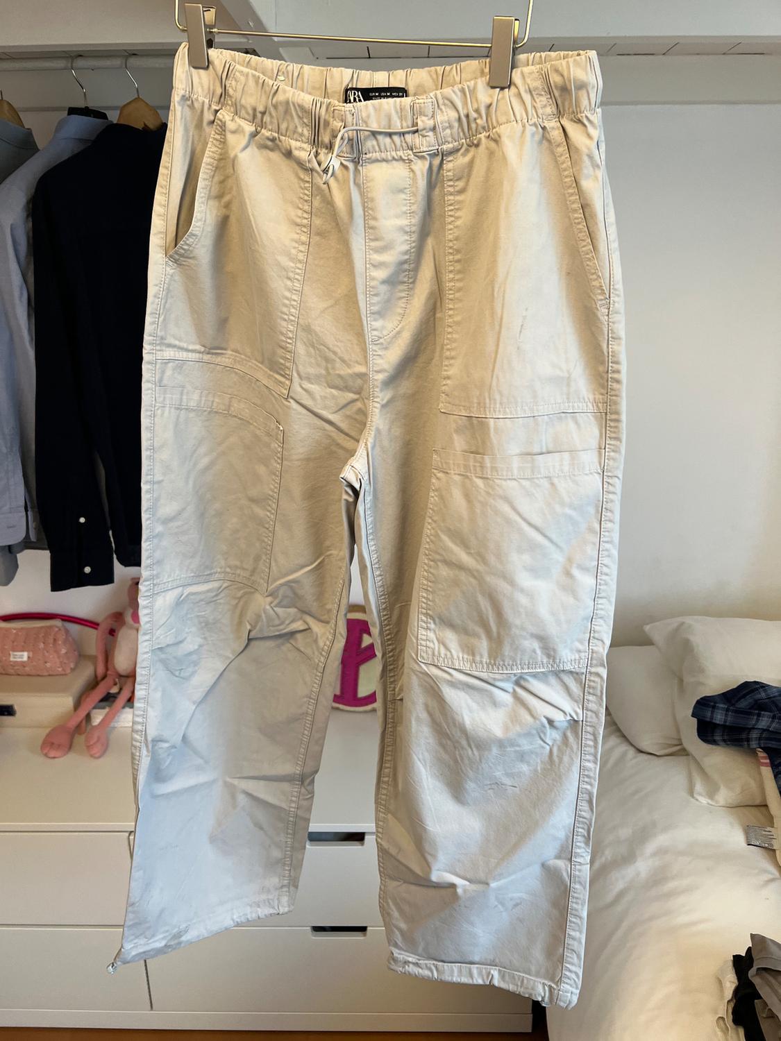 Beige trousers hanging on a hanger, with large pockets and an elasticated waist. Background with clothes hanging and a messy bed.