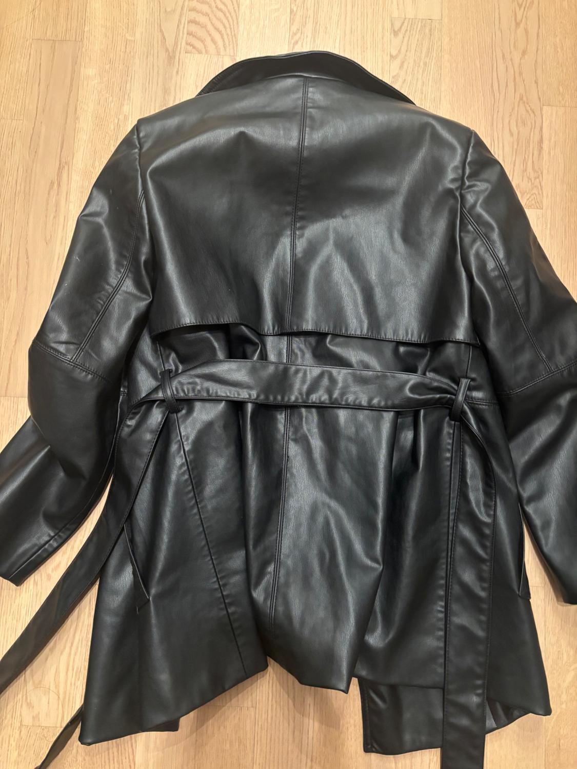 Black faux leather coat with belt, seen from behind, on a light wooden floor.