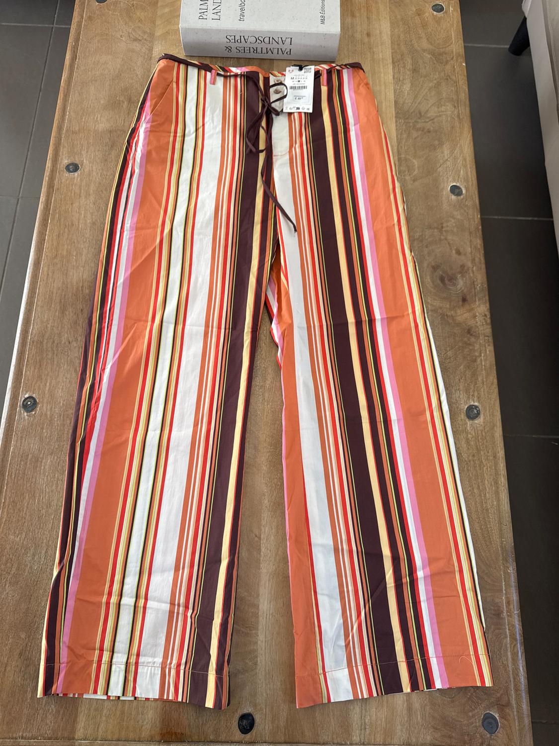 Vertical striped trousers in shades of orange, brown, and white on a wooden table. Label visible on the waistband.