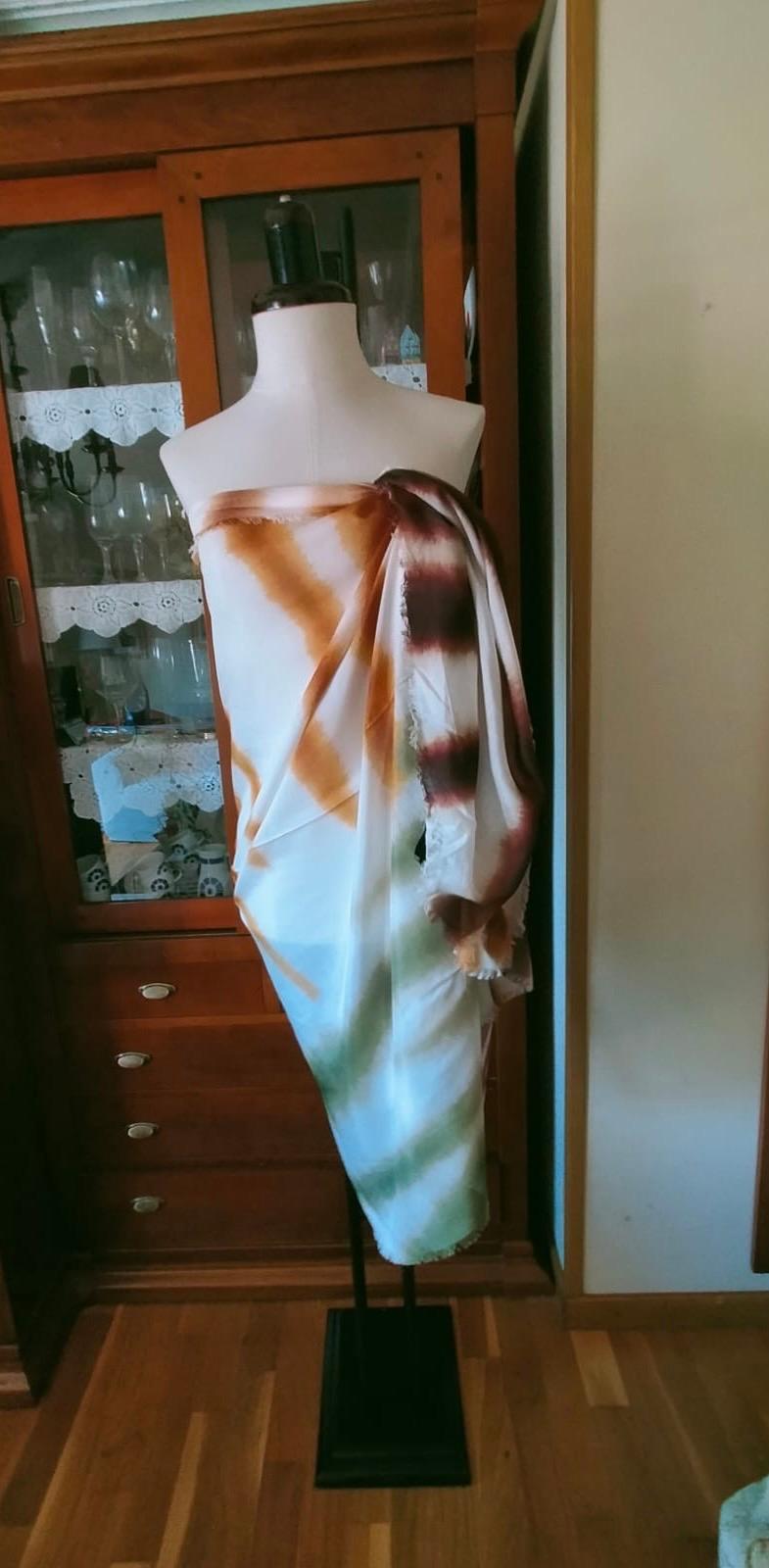 Mannequin with brown, orange, and green fabric, draped in a setting of wooden furniture and display cabinets with glasses.