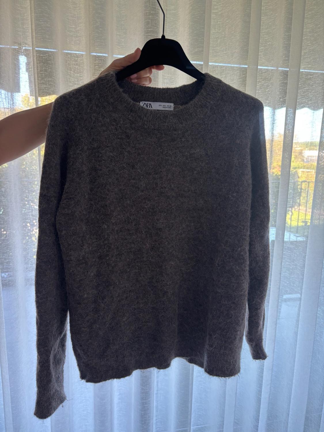 A hand holds a dark grey textured knit jumper on a black hanger in front of white translucent curtains. ZARA label.