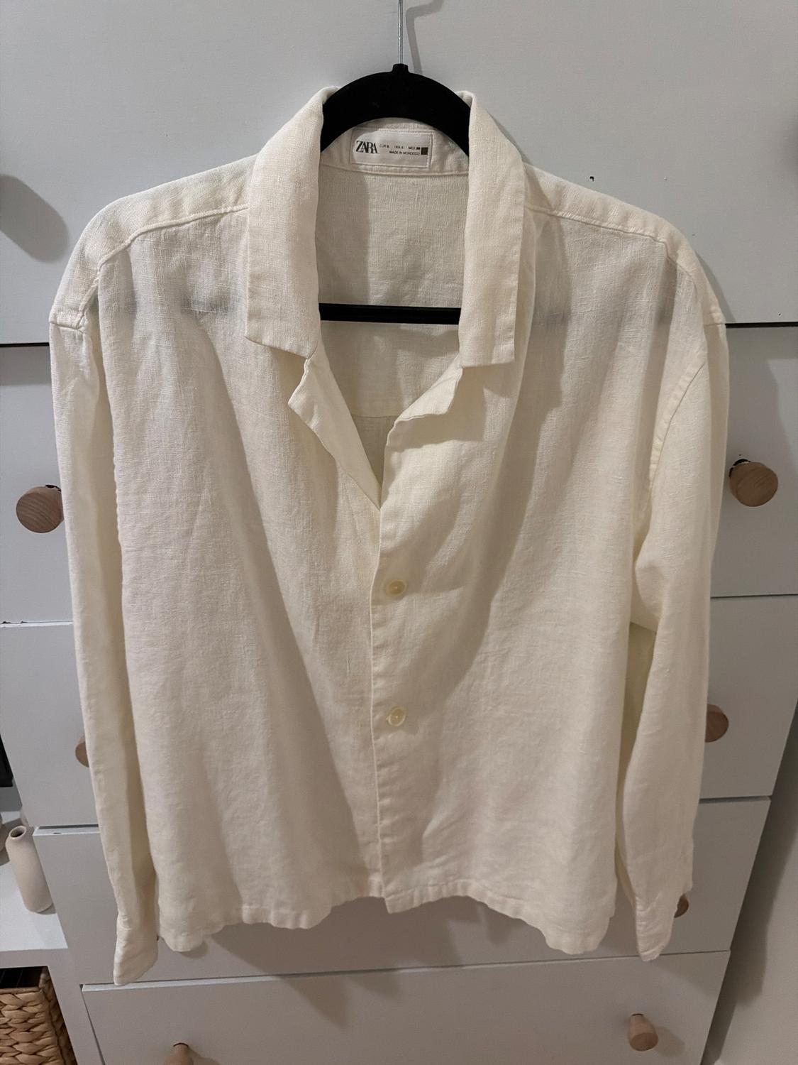 White linen shirt hanging on a black hanger in front of a white cabinet with wooden drawers. Label visible on the collar.