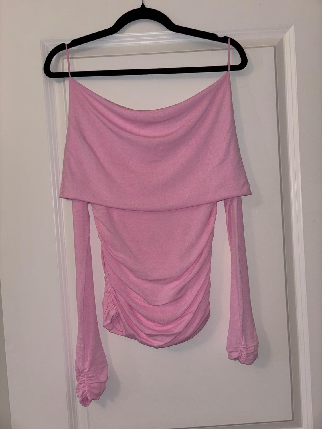 Pink, long-sleeved, off-the-shoulder blouse with ruching, hanging on a black coat hanger over a white door.