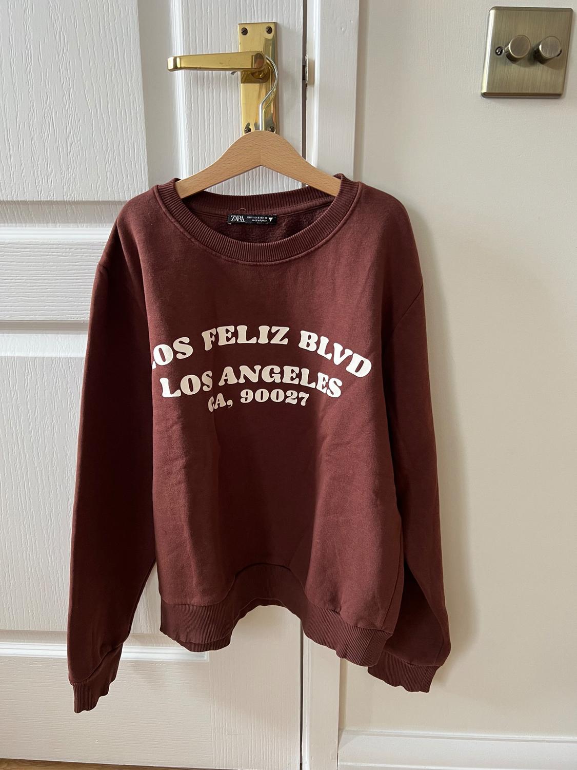 Brown sweatshirt hanging on a white door, with "LOS FELIZ BLVD LOS ANGELES CA. 90027" text in white letters.