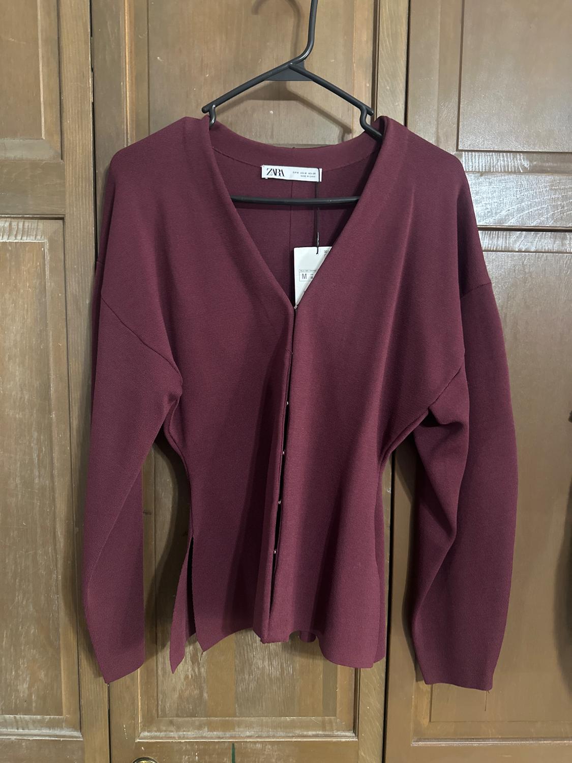Burgundy long-sleeved cardigan hanging on a black hanger, with front buttons and visible tag, against a wooden door background.