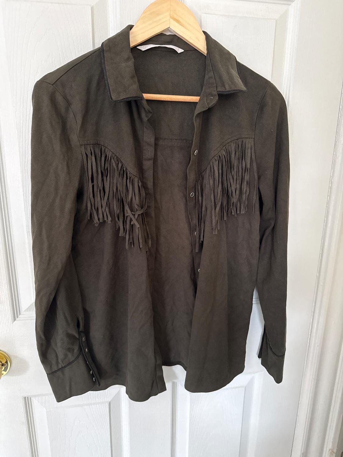 Brown jacket with fringe on the chest, hanging on a wooden hanger in front of a white door.