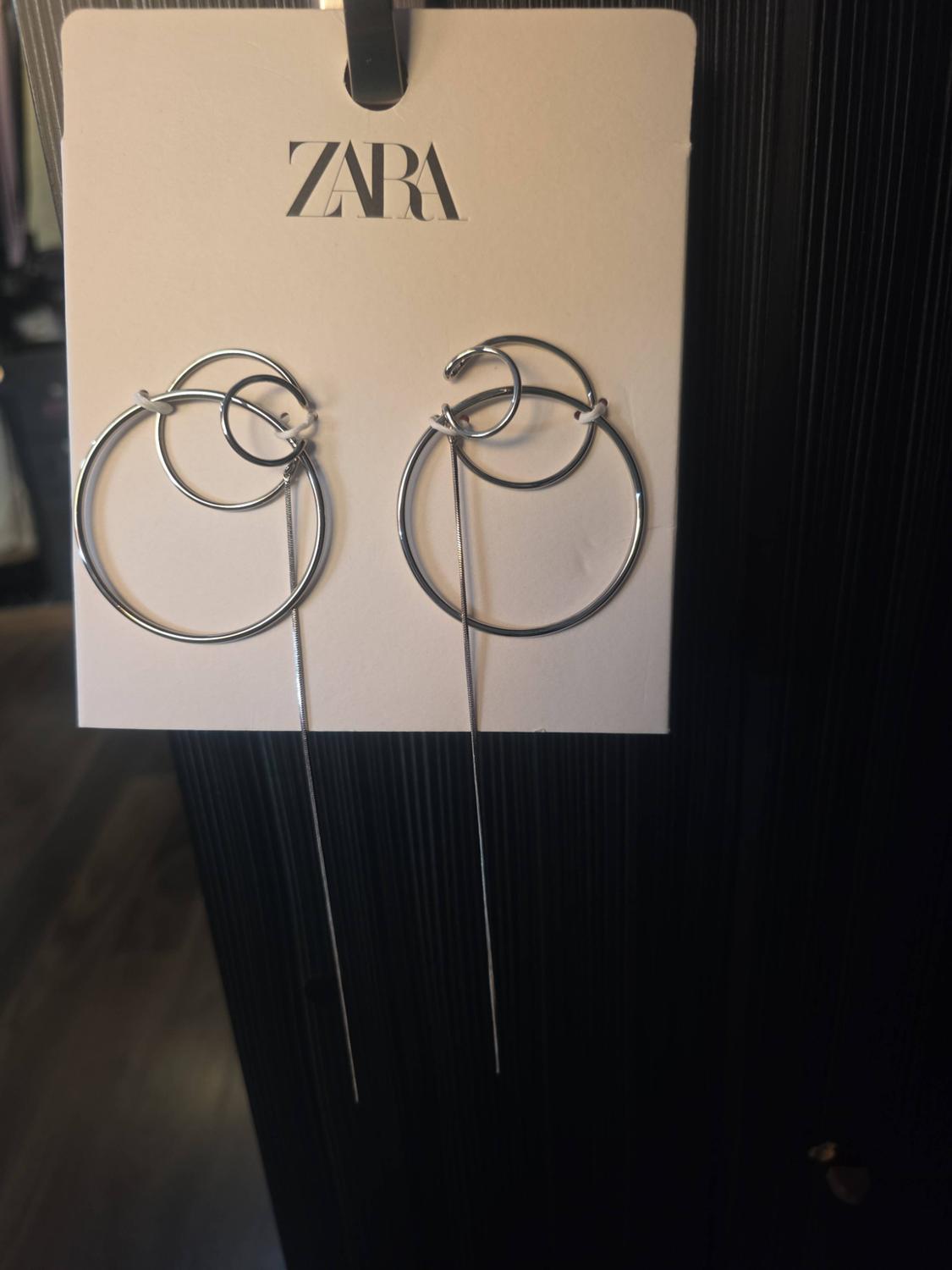 Silver-tone interlocking hoop earrings with a hanging chain on a white display card with ZARA text, on a dark background.