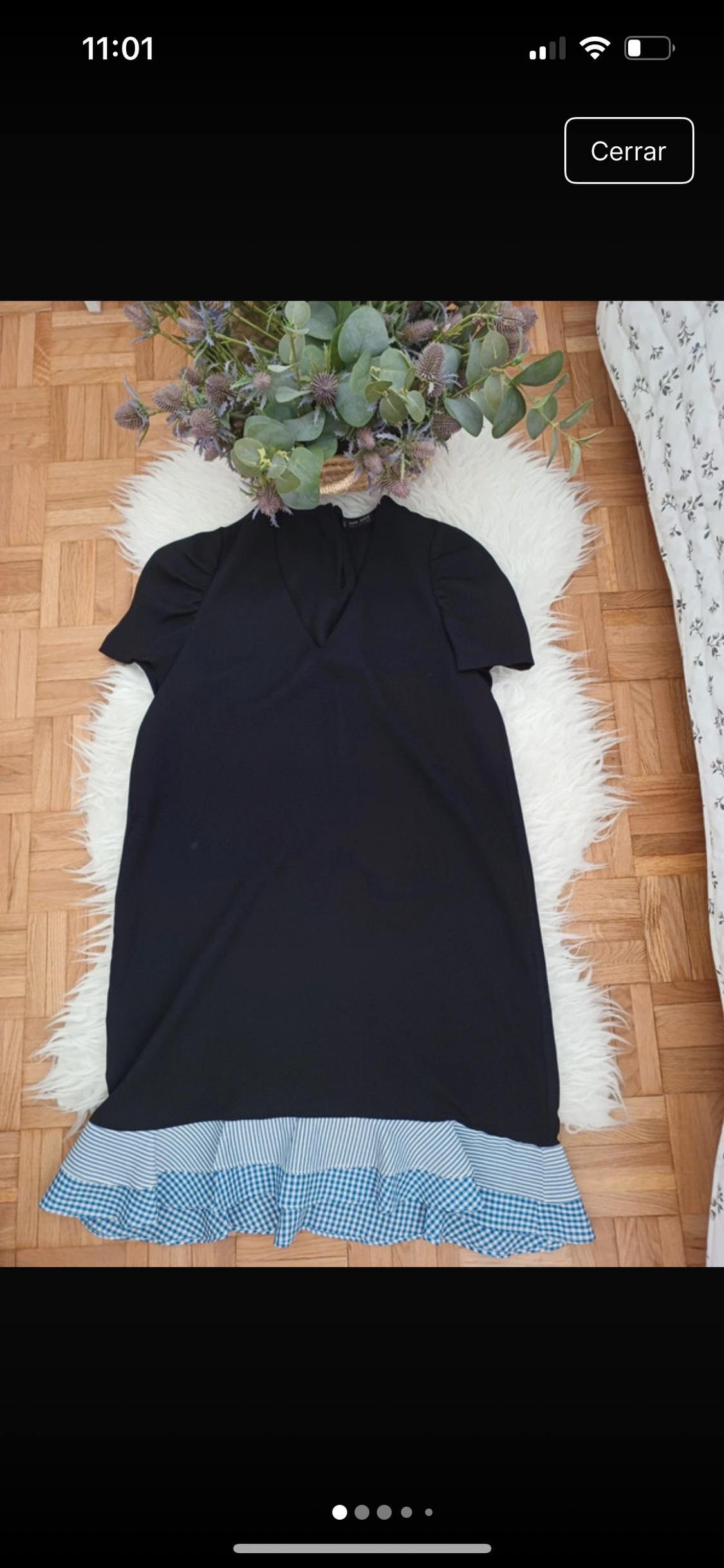 Μαύρο φόρεμα με κοντά μανίκια και μπλε καρό φούστα πάνω σε λευκό χαλί. Φόντο παρκέ και διακοσμητικά φυτά.