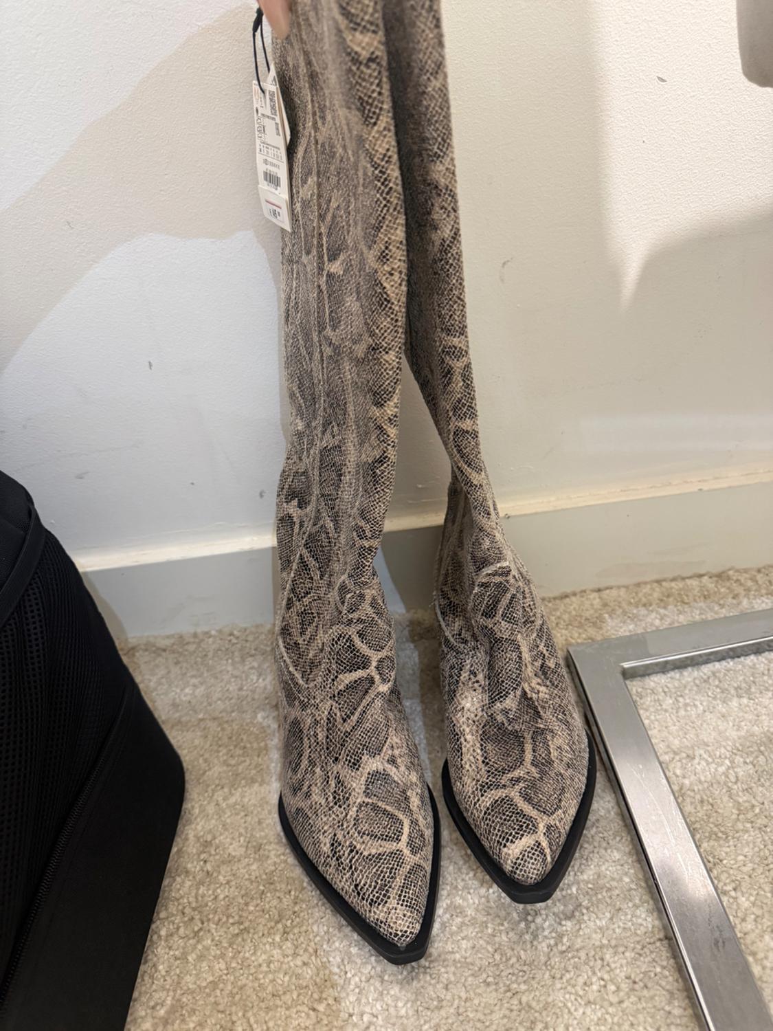 High boots with snake print in beige and black tones, on a light carpet. Visible tag hanging from one boot.